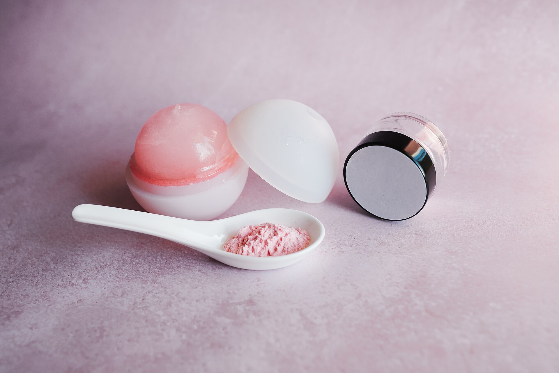 Two cosmetic jars and a small spoon of pink powder arranged on a pale pink surface